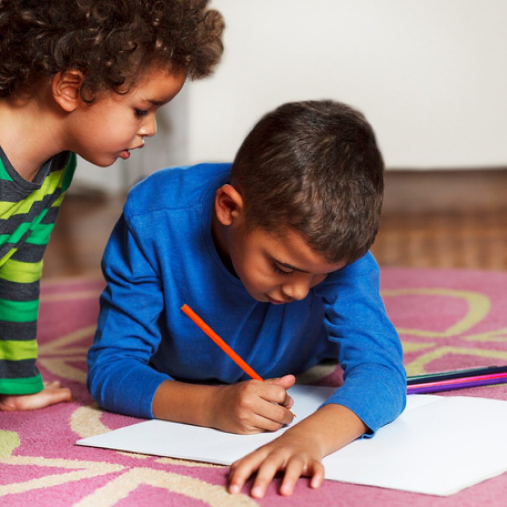 Cute school boys lying on the carpet, at home, drawing with crayons.