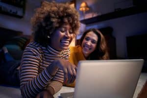 Two cheerful multi-ethnic women looking at computer lying on carpet in dark living room.